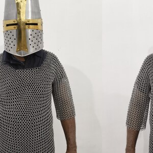 May include: A silver chainmail shirt with a short sleeve design. The shirt is worn by a person wearing a silver helmet with a gold cross.