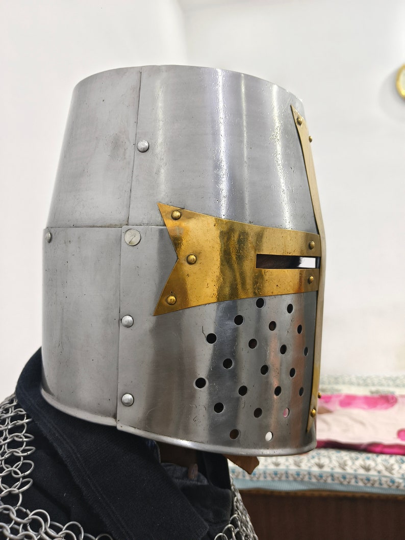 May include: A silver knight helmet with a brass cross on the front. The helmet has a visor with small holes for ventilation.