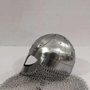 May include: A silver helmet with a chainmail coif. The helmet has a rounded top and a face mask with a small eye opening. The chainmail coif is attached to the helmet and hangs down to cover the neck.