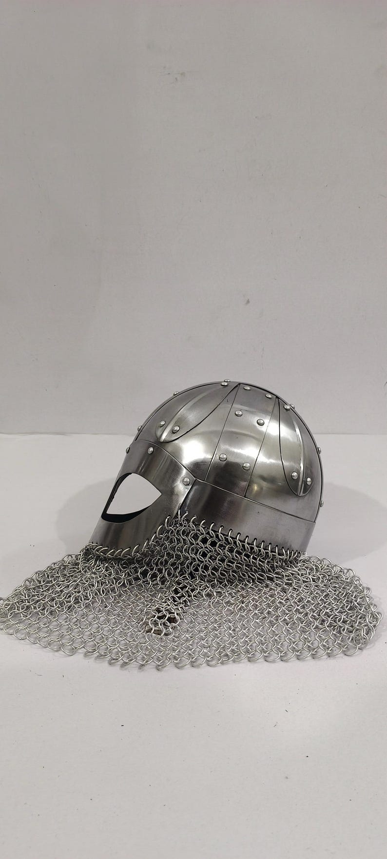 May include: A silver helmet with a chainmail coif. The helmet has a rounded top and a face mask with a small eye opening. The chainmail coif is attached to the helmet and hangs down to cover the neck.
