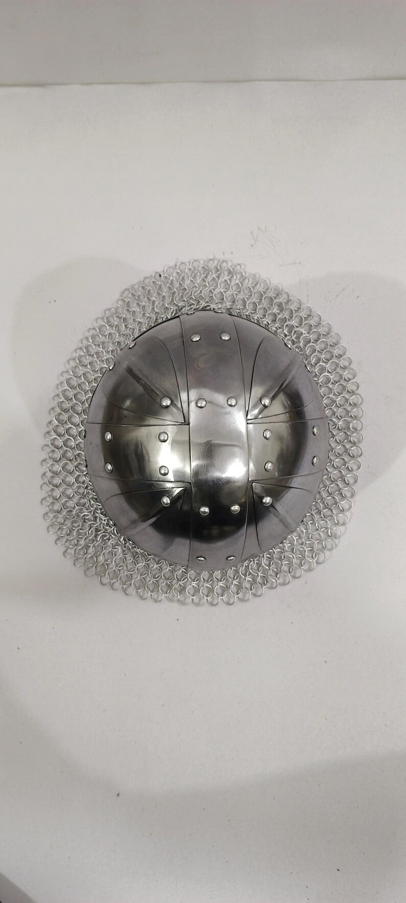 May include: A silver helmet with a chainmail coif. The helmet has a rounded dome shape with a series of raised ridges and rivets.
