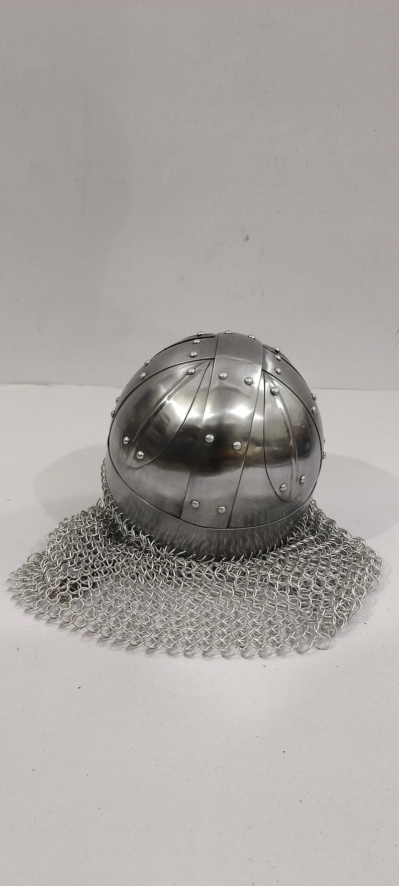May include: A silver helmet with a chainmail coif. The helmet is made of metal plates and has a rounded shape. The chainmail coif is made of small metal rings.