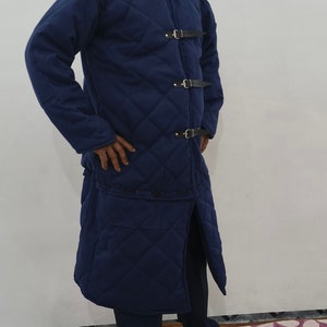 May include: A long, quilted, navy blue coat with a stand-up collar and four leather straps with buckles. The coat has a long, straight cut and a slit in the back.