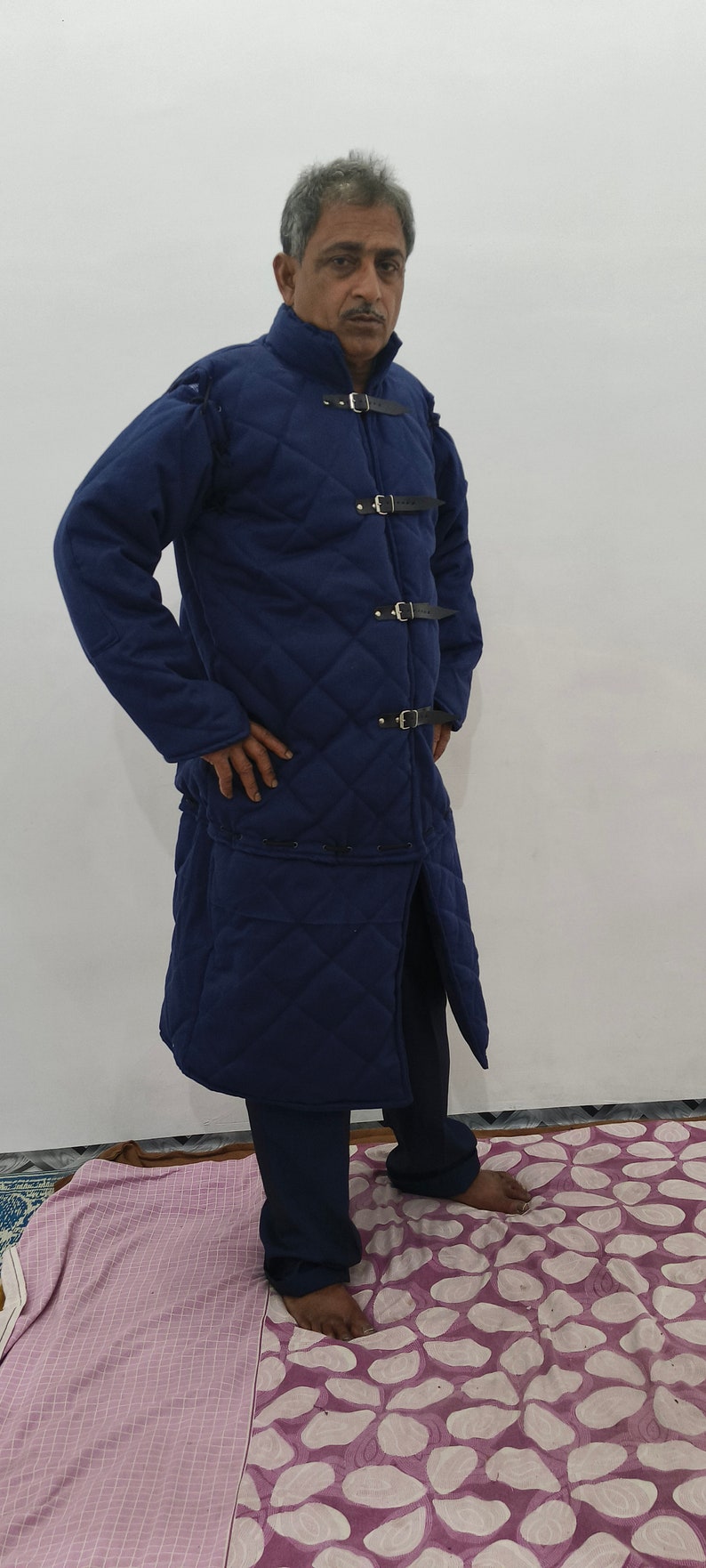 May include: A long, quilted, navy blue coat with a stand-up collar and four leather straps with buckles. The coat has a long, straight cut and a slit in the back.