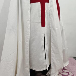 Op de afbeelding: Een witte robe met een rood kruis op de borst. De robe wordt gedragen door een persoon die op een vloer met een patroon staat.