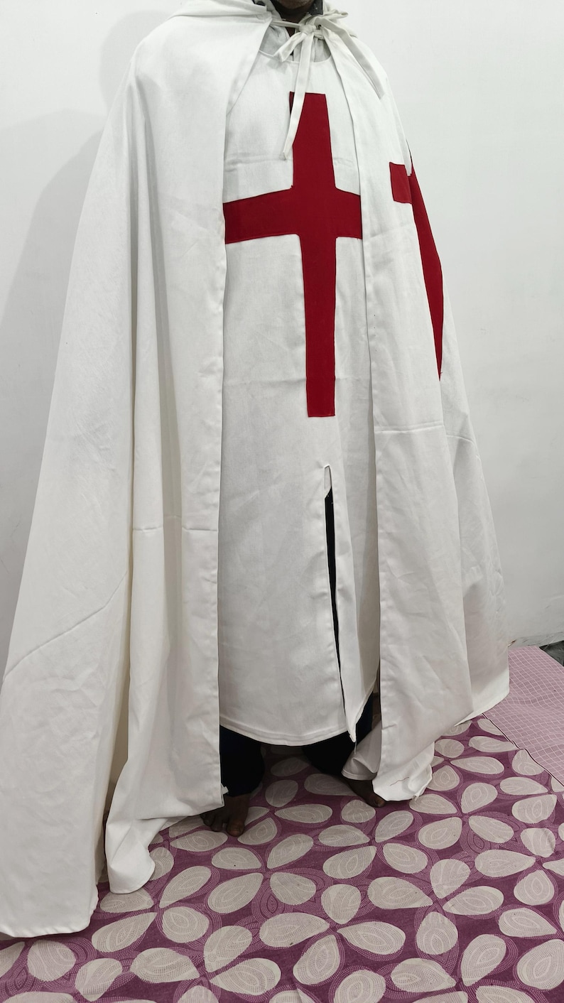 Op de afbeelding: Een witte robe met een rood kruis op de borst. De robe wordt gedragen door een persoon die op een vloer met een patroon staat.