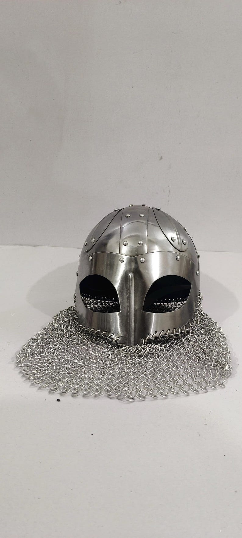 May include: A silver helmet with a chainmail coif. The helmet has a rounded top with a series of raised ridges and is decorated with rivets. The chainmail coif is made of small metal rings linked together.