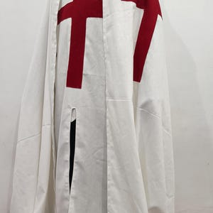 Op de afbeelding: Een witte kapmantel met een rood kruis op de voorkant. De mantel is gemaakt van een zachte, lichte stof en heeft een lang, vloeiend ontwerp.