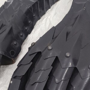 May include: A pair of black metal gauntlets with a scaly design. The gauntlets are made of metal plates that are riveted together. The gauntlets are designed to protect the hands and fingers during combat.