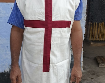 Peut inclure: Un homme portant une tunique blanche avec une croix rouge sur le devant. Il se tient devant un mur bleu et une porte en bois.