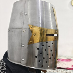 May include: A silver knight helmet with a brass cross on the front. The helmet has a visor with small holes for ventilation.