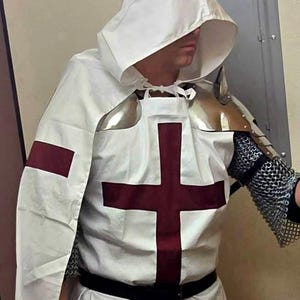 Op de afbeelding: Een witte tuniek met capuchon en een rood kruis op de borst. De tuniek wordt gedragen over een kettingharnas en heeft een zwarte riem.