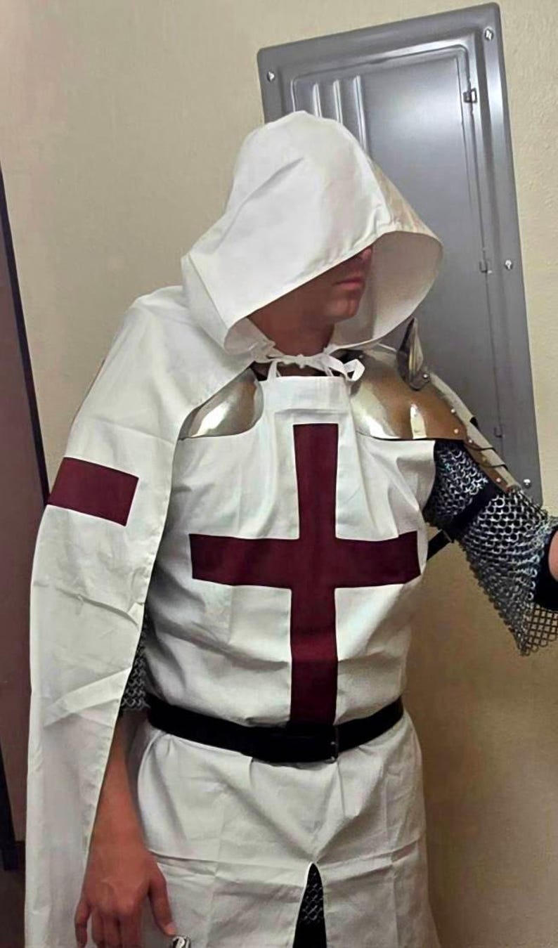Op de afbeelding: Een witte tuniek met capuchon en een rood kruis op de borst. De tuniek wordt gedragen over een kettingharnas en heeft een zwarte riem.