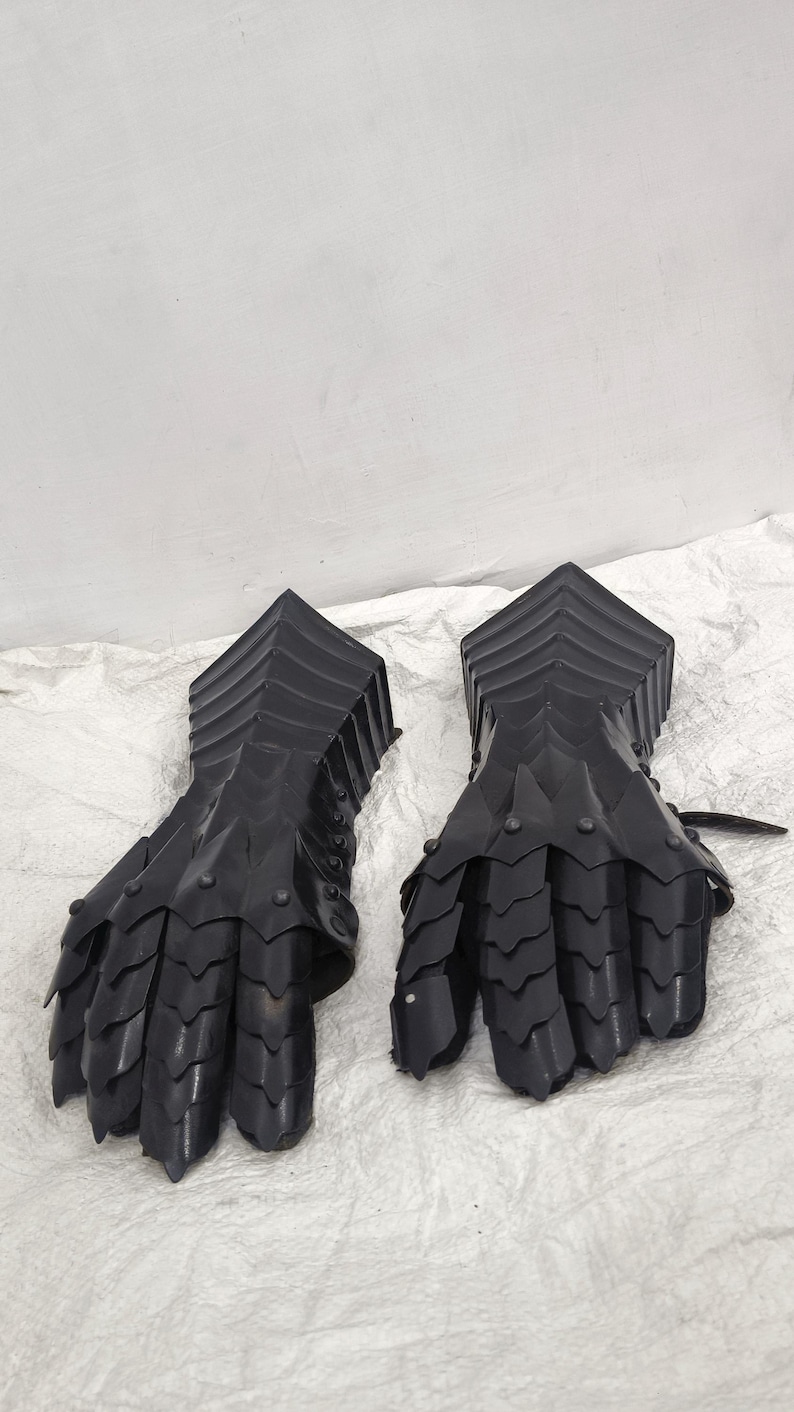 May include: A pair of black metal gauntlets with a scaly design. The gauntlets are made of interlocking metal plates and have a pointed tip on each finger.