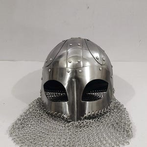 May include: A silver Viking helmet with a chainmail coif. The helmet has a rounded top with a pointed nose guard and eye holes. The chainmail coif is attached to the helmet and covers the neck and shoulders.