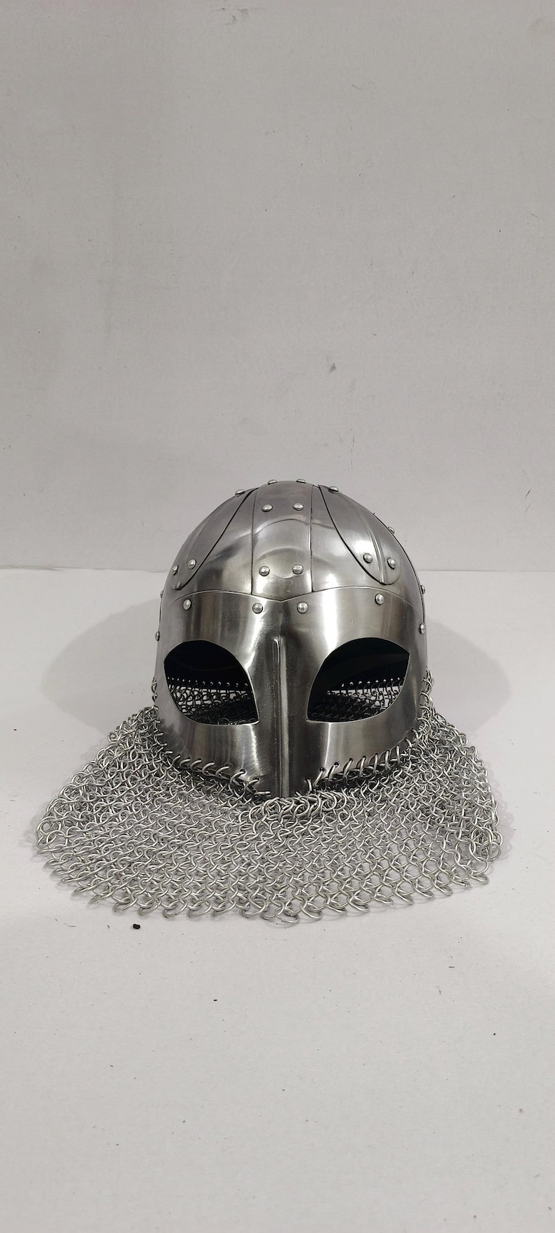 May include: A silver Viking helmet with a chainmail coif. The helmet has a rounded top with a pointed nose guard and eye holes. The chainmail coif is attached to the helmet and covers the neck and shoulders.