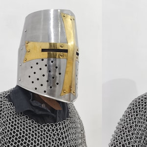 May include: A silver helmet with a gold cross design. The helmet has a mesh design and is worn by a person wearing chainmail.