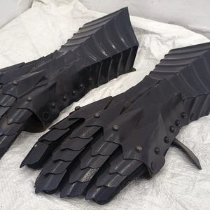May include: A pair of black metal gauntlets with a scaly design. The gauntlets are made of metal plates that are connected by rivets. The gauntlets are designed to protect the hands and fingers during combat.