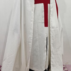 Op de afbeelding: Een witte kapmantel met een rood kruis op de voorkant. De mantel wordt gedragen door een persoon, maar de persoon is niet zichtbaar op de afbeelding.