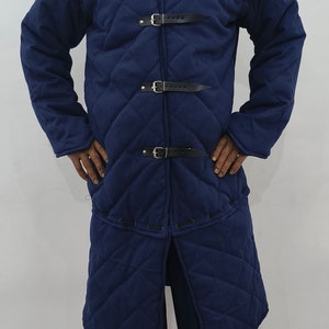 May include: A long, dark blue quilted coat with a stand-up collar and multiple leather straps and buckles. The coat is worn by a person standing on a patterned rug.