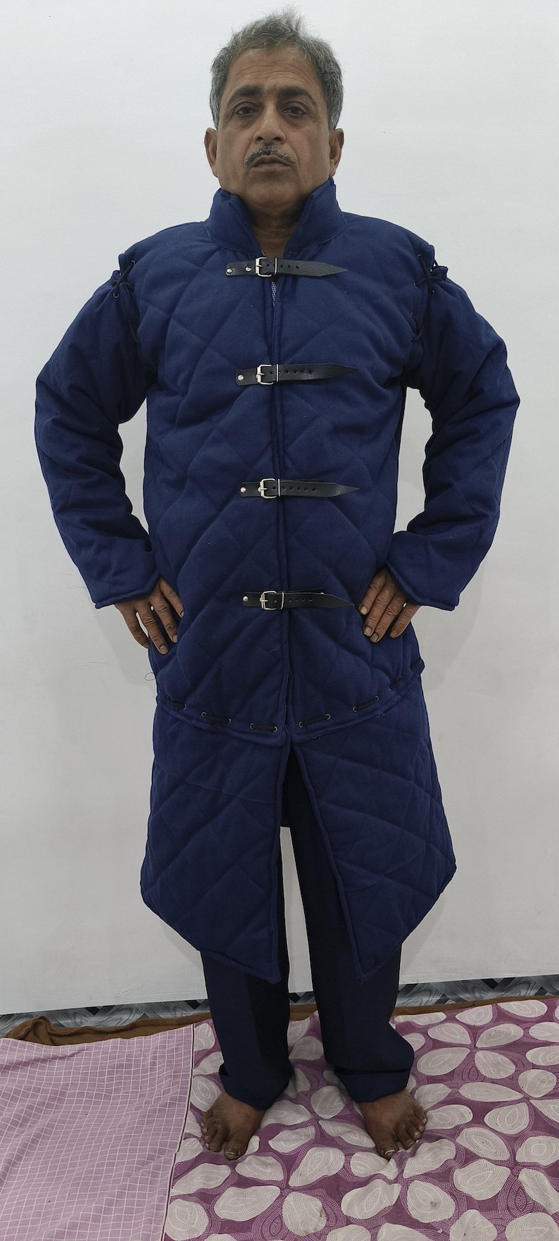 May include: A long, dark blue quilted coat with a stand-up collar and multiple leather straps and buckles. The coat is worn by a person standing on a patterned rug.