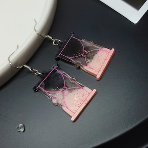 Puede incluir: Un par de pendientes de reloj de arena negro y rosa. Los pendientes están hechos de resina y tienen un gancho plateado.