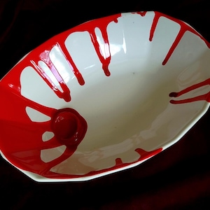 Puede incluir: Cuenco de cerámica blanco de forma ovalada con un diseño de pintura roja goteando alrededor del borde y el interior. El cuenco tiene una pequeña hendidura circular en un lado. La pintura roja crea un contraste llamativo.