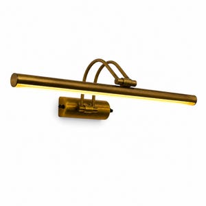 May include: A wall-mounted picture light with an antique brass finish. The fixture features a long, horizontal cylindrical lamp head with an integrated LED light strip, connected by two curved metal arms to a rectangular mounting base plate.