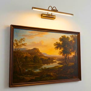 May include: A rectangular landscape painting in a dark brown frame featuring a river valley at sunset with golden light, trees, and distant hills. Above the painting is a horizontal brass picture light fixture emitting a warm glow.