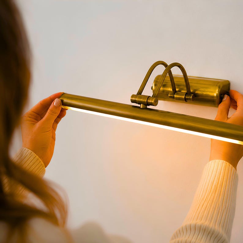 May include: A brass-toned picture light with a cylindrical lamp head and a rectangular wall-mounting base. The fixture features two curved support arms connecting the light bar to the base, emitting a warm, bright glow from the underside.