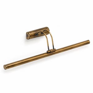 Op de afbeelding: Een wandlamp voor schilderijen met een antieke messing afwerking. Het armatuur is voorzien van een rechthoekige basisplaat, twee gebogen steunarmen en een lange, slanke horizontale cilindrische lichtbalk die uitsteekt vanaf de muur.