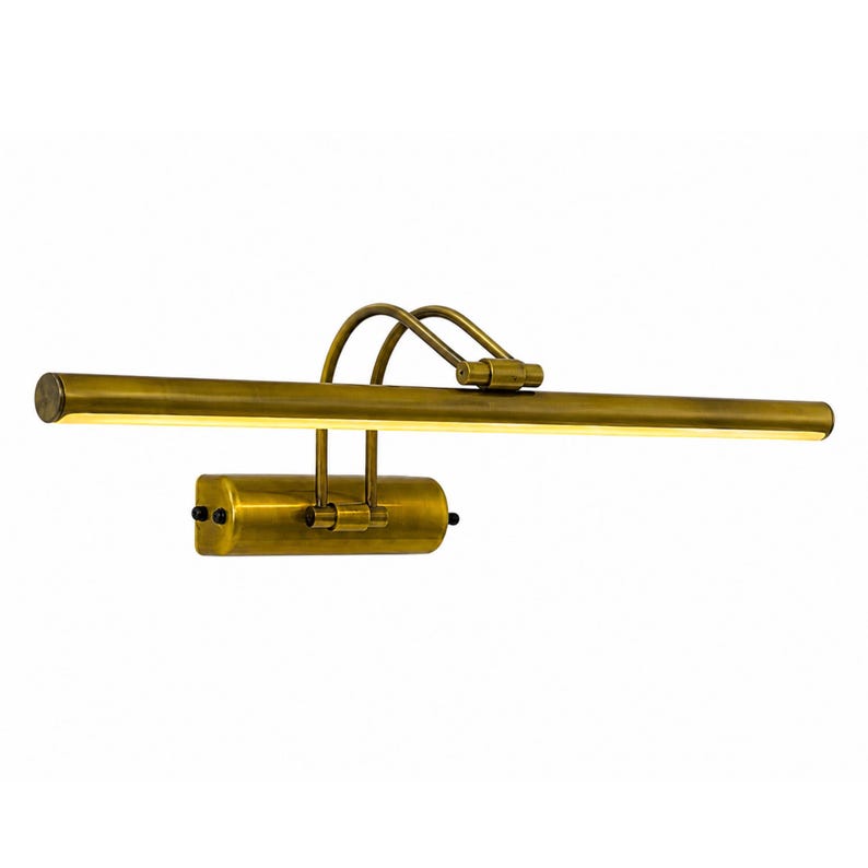 May include: A brass picture light with a long, horizontal cylindrical lamp head and a smaller, rectangular base. The fixture features two curved arms connecting the base to the lamp, with a warm, illuminated light strip along the front edge.