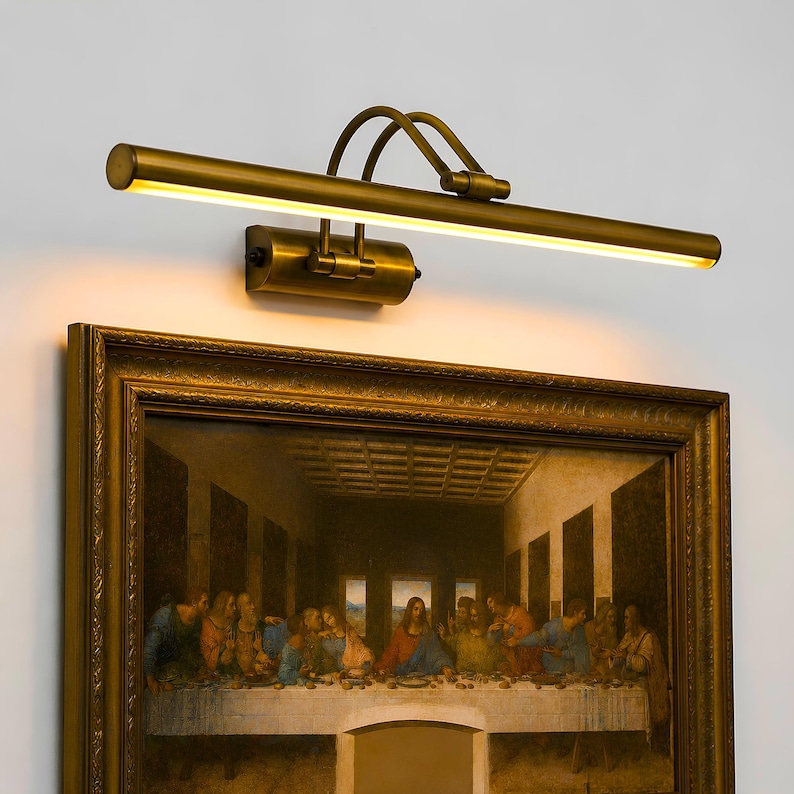 May include: A brass picture light fixture with a long, cylindrical horizontal bar and two curved support arms, mounted above a framed painting of The Last Supper. The light emits a warm, downward glow, illuminating the top of the ornate gold frame.