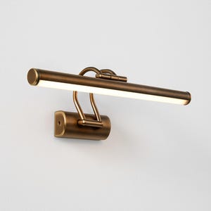 May include: A bronze-colored picture light fixture with a rectangular light bar. The light bar is mounted on curved supports and a rectangular base. The fixture is designed to be mounted on a wall.