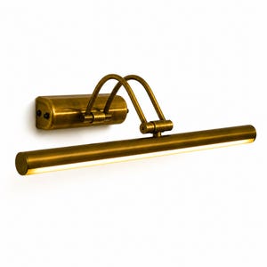 May include: A wall-mounted picture light with an antique brass finish. The fixture features a rectangular base, two curved support arms, and a long, cylindrical horizontal lamp head with an integrated LED light strip emitting a warm glow.