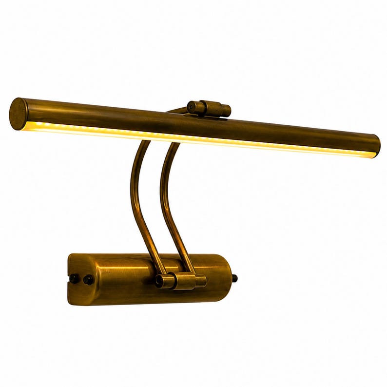 May include: A wall-mounted picture light with an antique brass finish. The fixture features a horizontal cylindrical lamp head with an integrated LED light strip, connected by two curved metal arms to a rectangular base with two side screws.