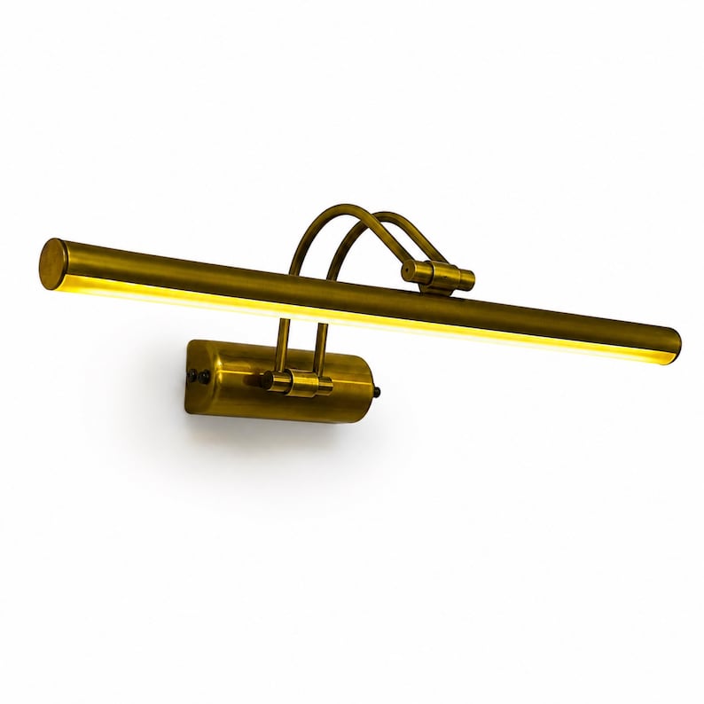 May include: A brass picture light with a long, cylindrical horizontal lamp head and a rectangular wall-mounting base. The fixture features two curved support arms connecting the base to the light, emitting a warm, soft glow from the underside.