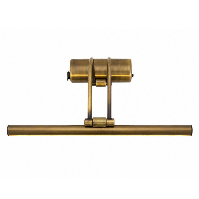 May include: An antique brass picture light featuring a horizontal tubular lamp head attached to a cylindrical wall-mounting base by two adjustable vertical arms. The metal finish has a brushed, weathered texture throughout the entire fixture.