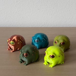 May include: Five small, round, 3D-printed frog figurines in various colors including metallic copper, teal, olive green, lime green, and muted green. Each frog features distinct, wide-set eyes with horizontal pupils and small textured bumps on their backs.