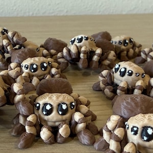 May include: A collection of small, three-dimensional printed spider figurines. Each spider features a tan body with dark brown accents on the legs and back, and large, black eyes with white highlights, creating a stylized, rounded appearance.