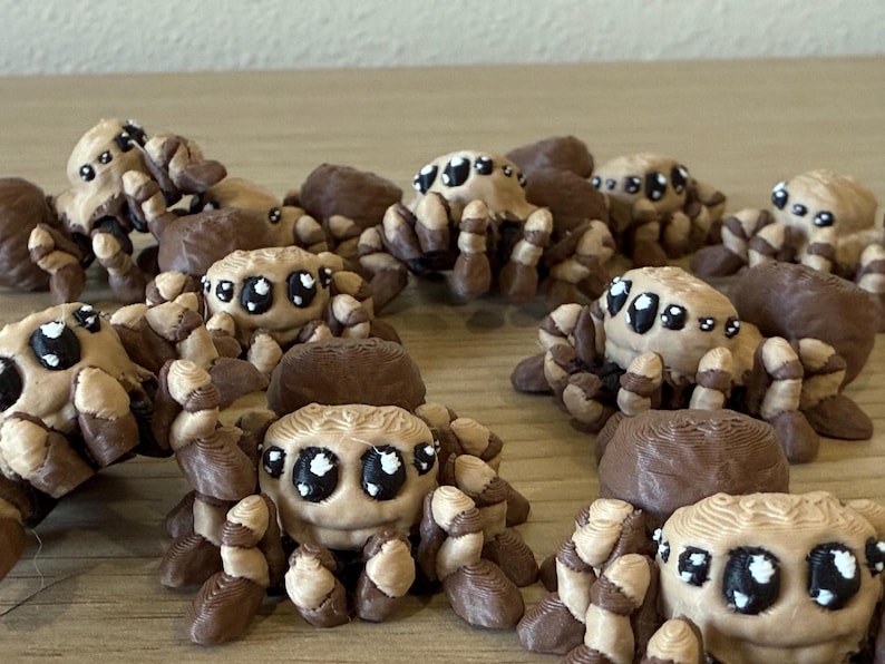 May include: A collection of small, three-dimensional printed spider figurines. Each spider features a tan body with dark brown accents on the legs and back, and large, black eyes with white highlights, creating a stylized, rounded appearance.