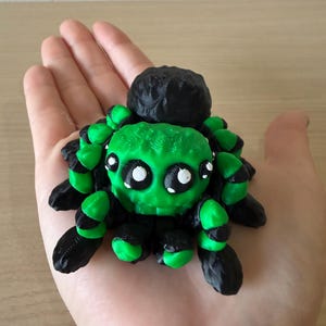 May include: A 3D printed spider figurine featuring a bright green head and body with black accents on the legs and abdomen. The spider has large, round, black eyes with white highlights, giving it a distinct appearance with textured surfaces.