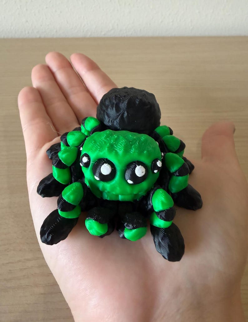 May include: A 3D printed spider figurine featuring a bright green head and body with black accents on the legs and abdomen. The spider has large, round, black eyes with white highlights, giving it a distinct appearance with textured surfaces.