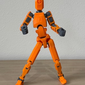 May include: An articulated action figure with a bright orange body and dark gray joints. The figure features a geometric head design, a chest plate embossed with the number 13, and clenched fists. It stands in a wide, dynamic pose.