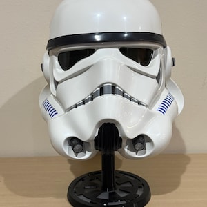 May include: A white Stormtrooper helmet with black eye lenses, a black brow trim, and blue accent stripes on the sides. The helmet is mounted on a black circular display stand with a central vertical support post and a textured base design.