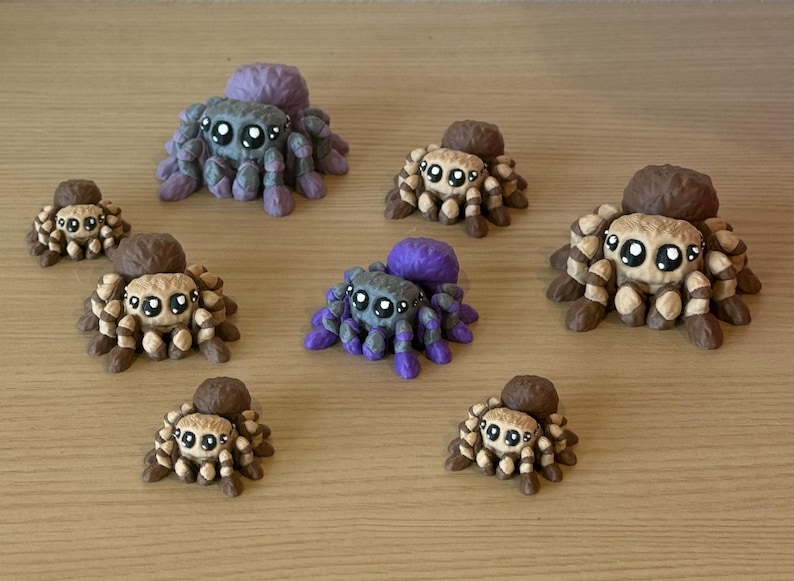 May include: A collection of seven small spider figurines arranged in a group. The spiders feature large, round eyes and are painted in two distinct color schemes: a purple and grey combination, and a brown and tan striped pattern.