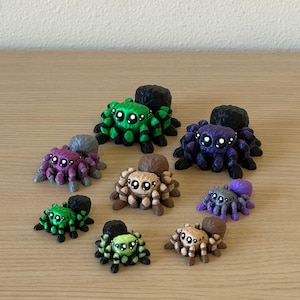 May include: A collection of eight small spider figurines in various colors including green, purple, brown, and black. Each spider features large, round, white-highlighted eyes and a rounded body shape, arranged in a cluster on a flat surface.