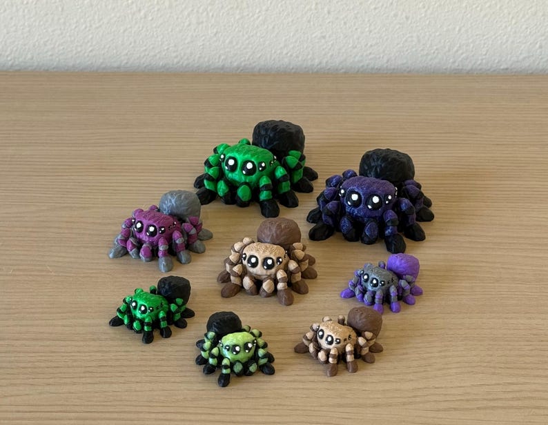May include: A collection of eight small spider figurines in various colors including green, purple, brown, and black. Each spider features large, round, white-highlighted eyes and a rounded body shape, arranged in a cluster on a flat surface.