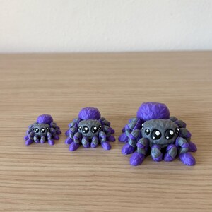 May include: Three small spider figurines in varying sizes, each featuring a textured purple body and a grey head with large, black, glossy eyes. The spiders have multiple purple and grey legs arranged around their bodies, creating a consistent appearance across all three items, which are displayed in a row from smallest to largest.
