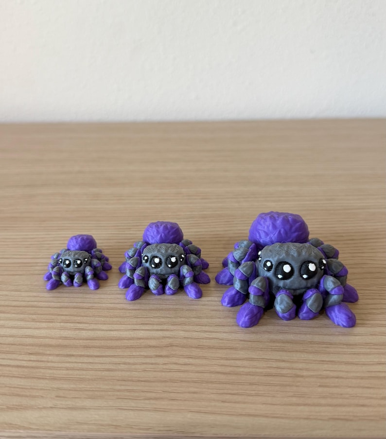 May include: Three small spider figurines in varying sizes, each featuring a textured purple body and a grey head with large, black, glossy eyes. The spiders have multiple purple and grey legs arranged around their bodies, creating a consistent appearance across all three items, which are displayed in a row from smallest to largest.